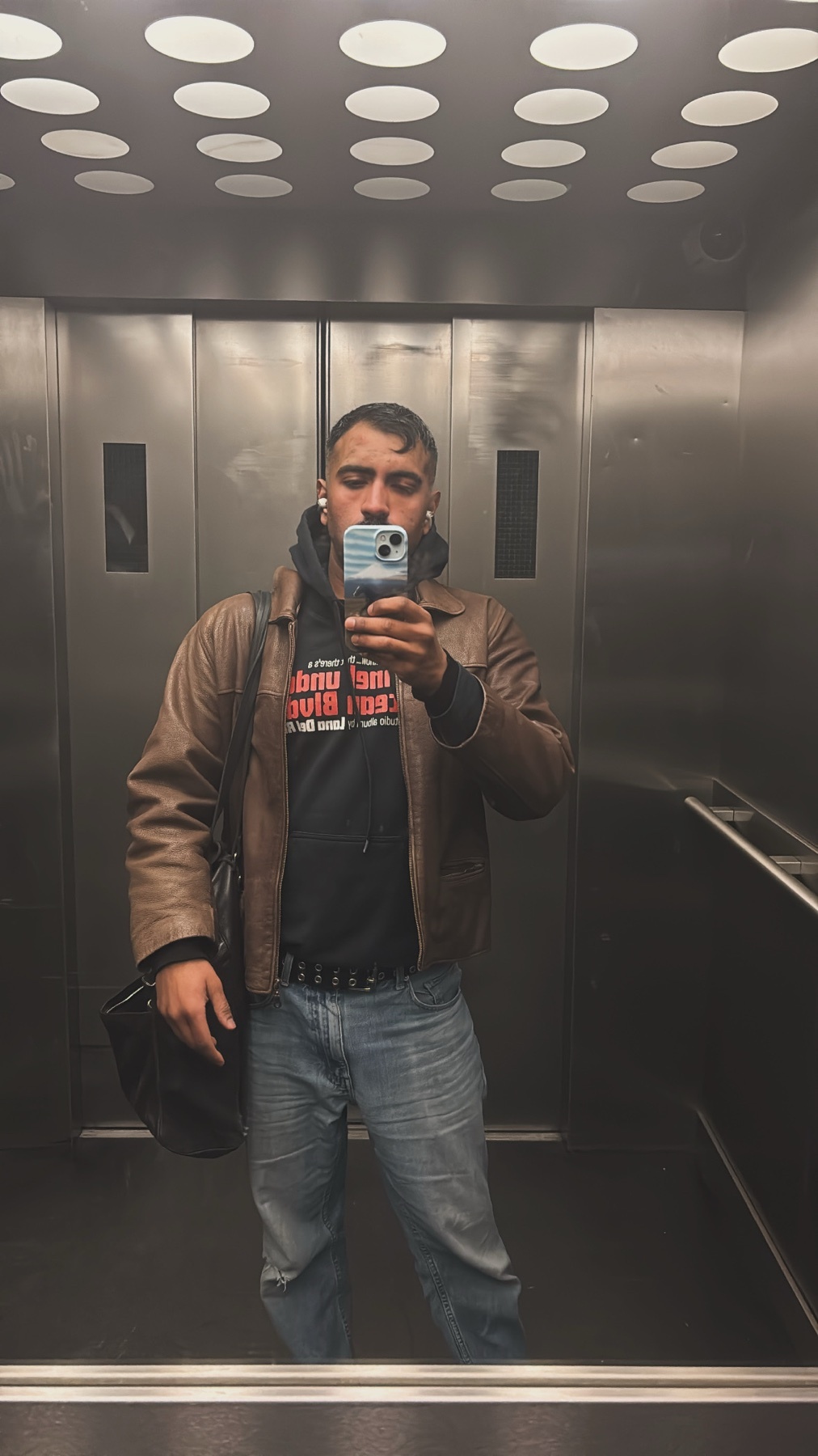A man takes a selfie in an elevator, wearing a brown leather jacket over a black hoodie and blue jeans. He is also carrying a black bag over his shoulder.