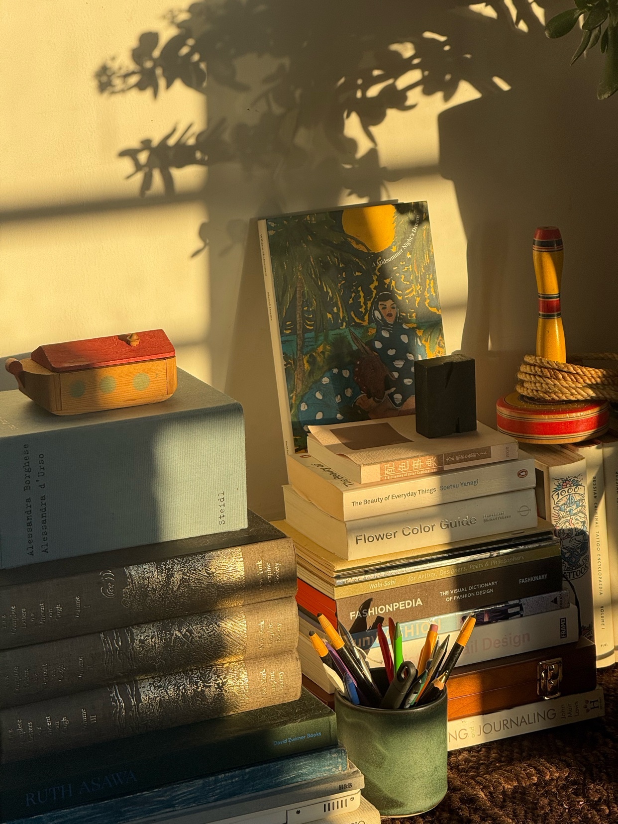 A stack of books and art pieces are illuminated by sunlight, creating a warm and inviting atmosphere. The composition highlights the beauty of everyday objects.