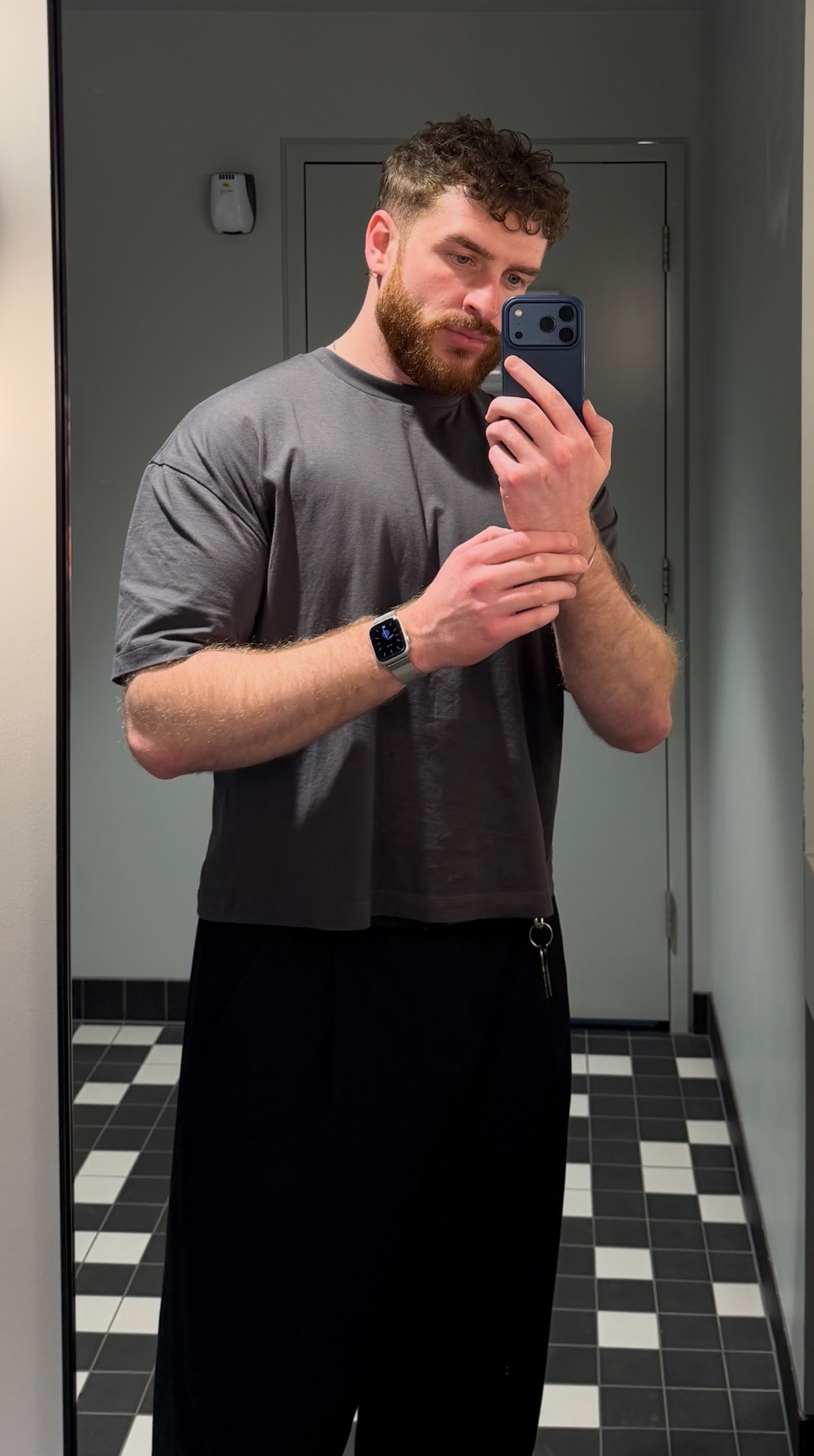A man takes a mirror selfie. He is wearing a gray t-shirt and black pants.