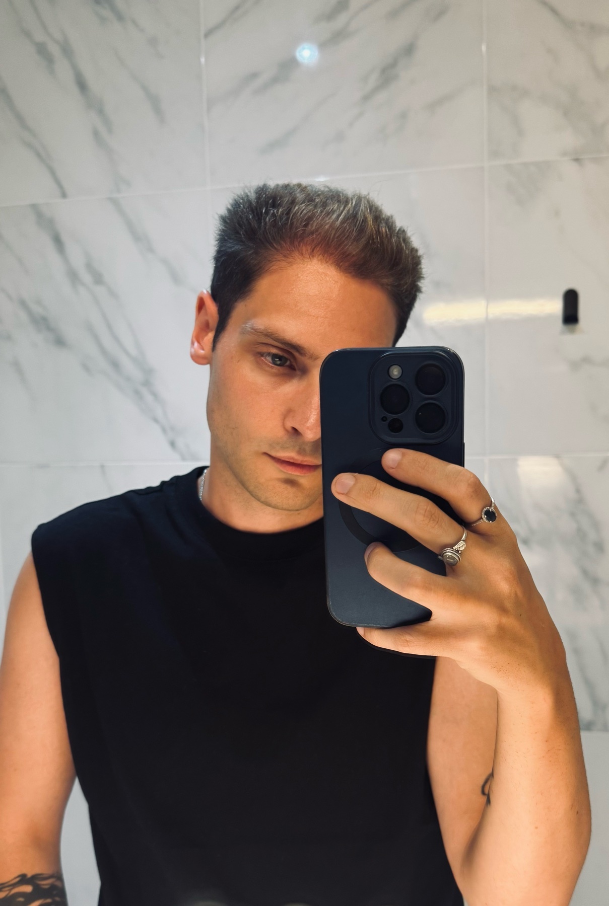 A man takes a selfie in a bathroom, holding his phone in front of his face. He is wearing a black sleeveless shirt and has short hair and tattoos on his arm.