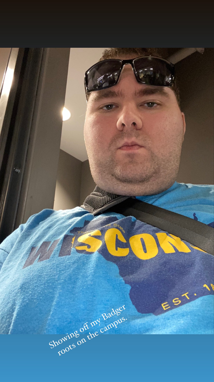 A man wearing sunglasses and a blue t-shirt with 'Wisconsin' printed on it poses for a selfie on campus.