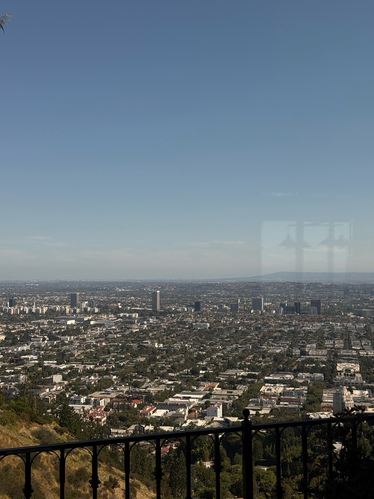 A sprawling cityscape stretches out under a clear blue sky, viewed from a high vantage point. Buildings of various sizes dot the landscape, with a railing in the foreground suggesting a viewpoint or balcony.