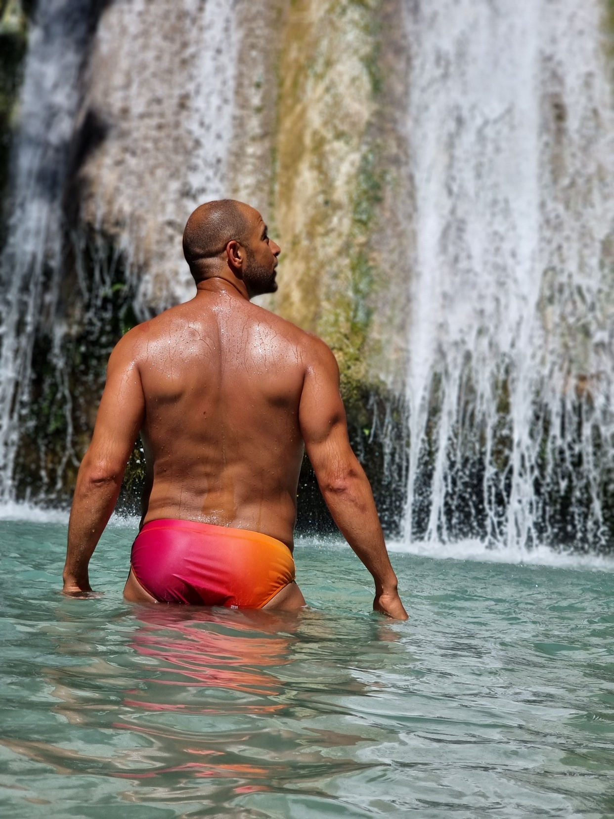 A muscular man stands in the water in front of a waterfall, wearing colorful swimming trunks. The water is clear and turquoise, and the waterfall cascades down a rocky cliff face.
