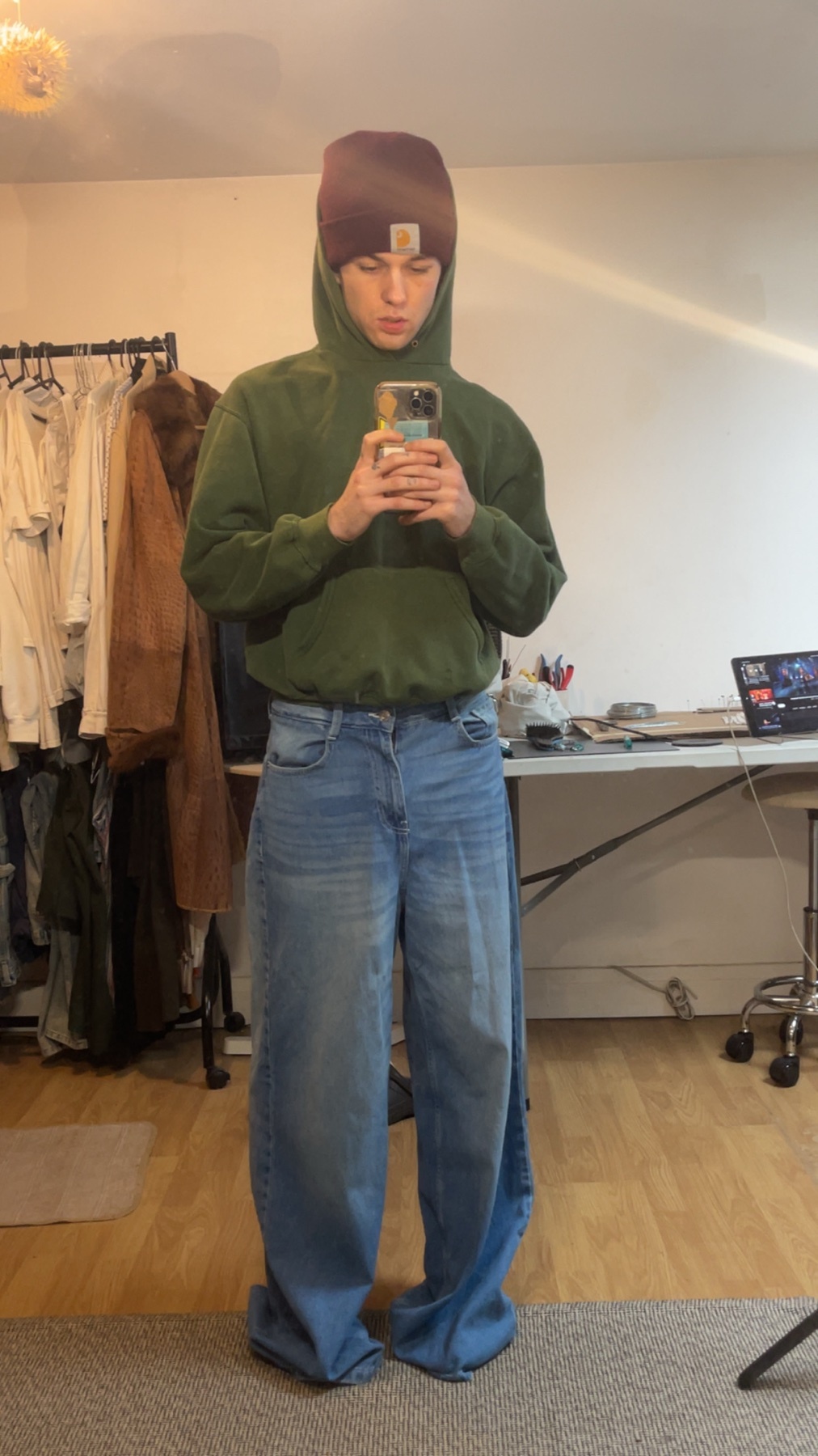 A man takes a mirror selfie, showcasing his outfit. He wears a green hoodie, oversized jeans, and a beanie.