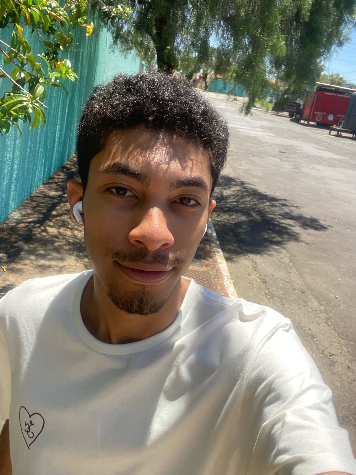 A young man with curly hair and stubble takes a selfie outdoors while wearing earbuds and a t-shirt.