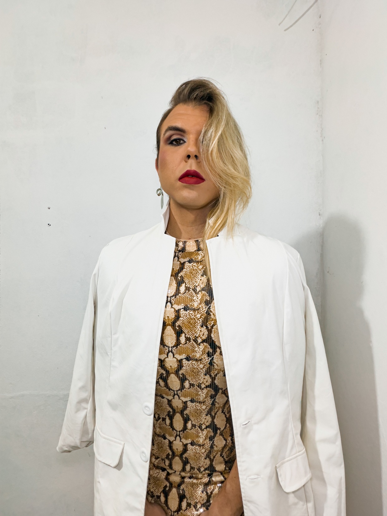 A drag queen poses in a white blazer over a snakeskin-print bodysuit. She wears bold red lipstick and has blonde hair styled to one side.