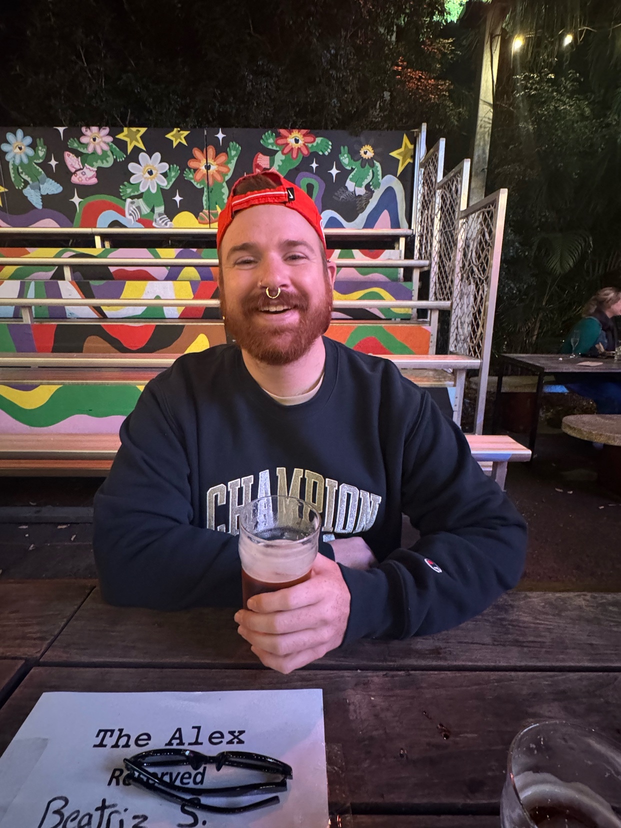 A man with a red beard and nose ring smiles at the camera while holding a glass of beer. He is wearing a red baseball cap and a dark sweatshirt with 'Champion' written on it. The background features a colorful mural and outdoor seating.