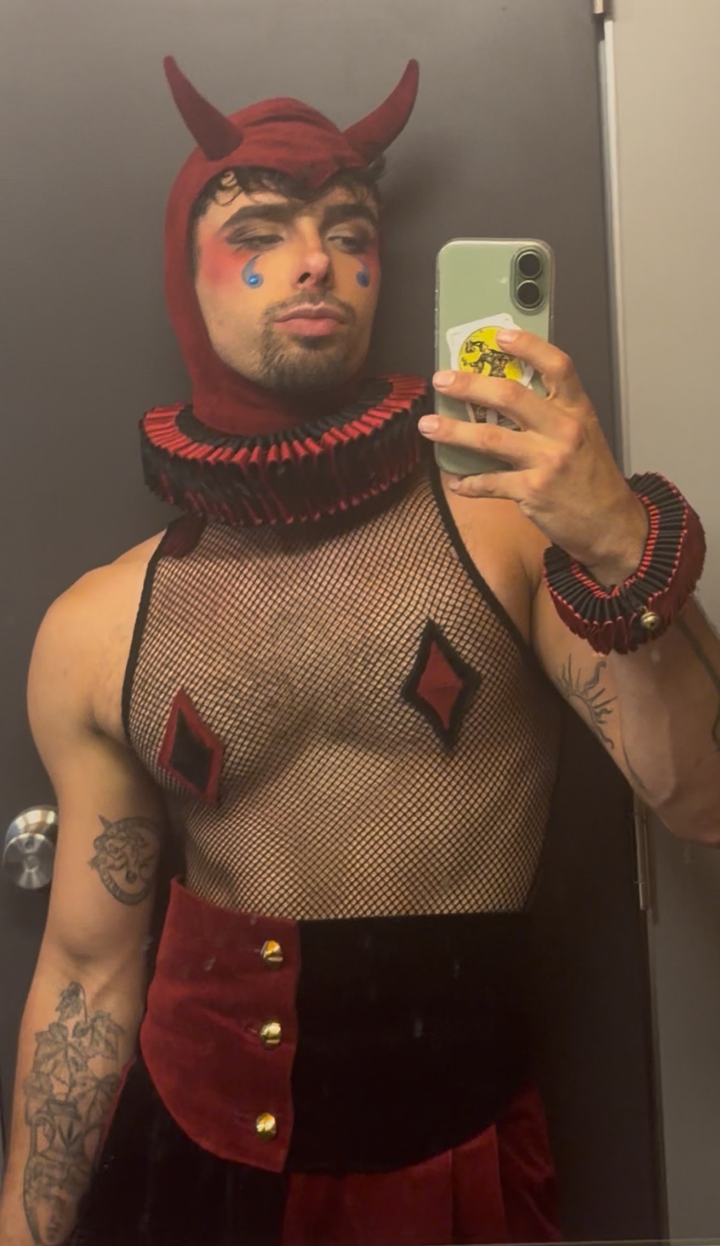 A man in a devil costume takes a selfie. The costume includes a red hood with horns, a ruffled collar, and a fishnet top with diamond accents.