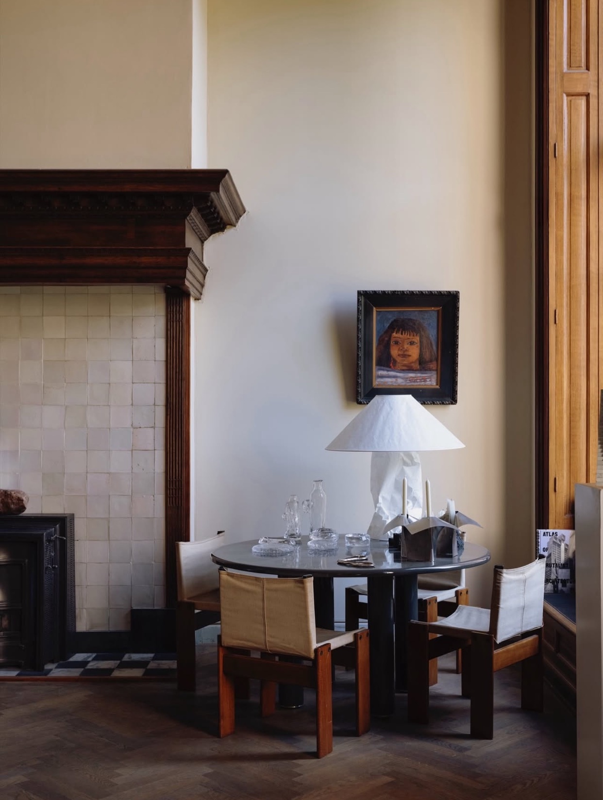 An interior shot featuring a round table with chairs, a lamp, and a painting on the wall. The room has a vintage aesthetic.