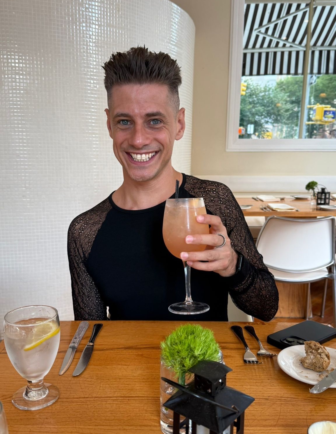 A man smiles while holding a cocktail at a restaurant. He is wearing a black shirt and has short, curly hair.
