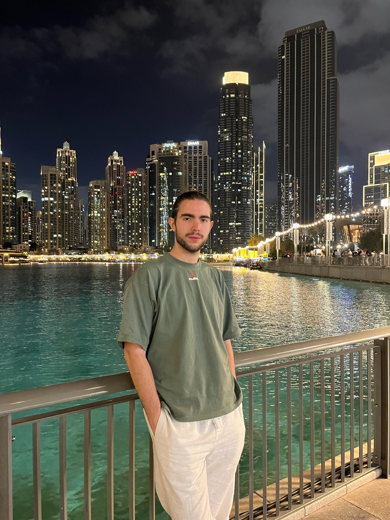 A man stands on a railing overlooking a body of water with a city skyline at night in the background. The skyscrapers are illuminated, reflecting on the water.