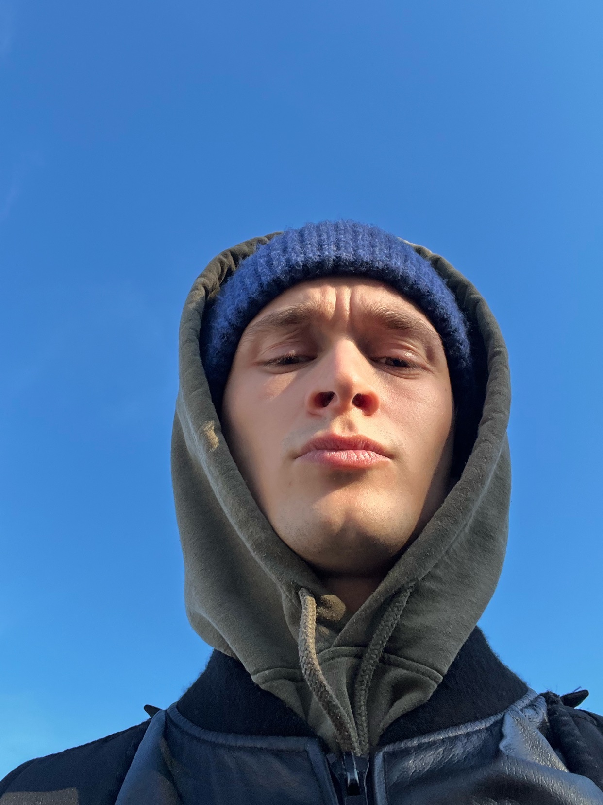 A man in a hoodie and beanie poses for a selfie against a bright blue sky. The photo captures a moment of happiness and appreciation for the sunny weather.