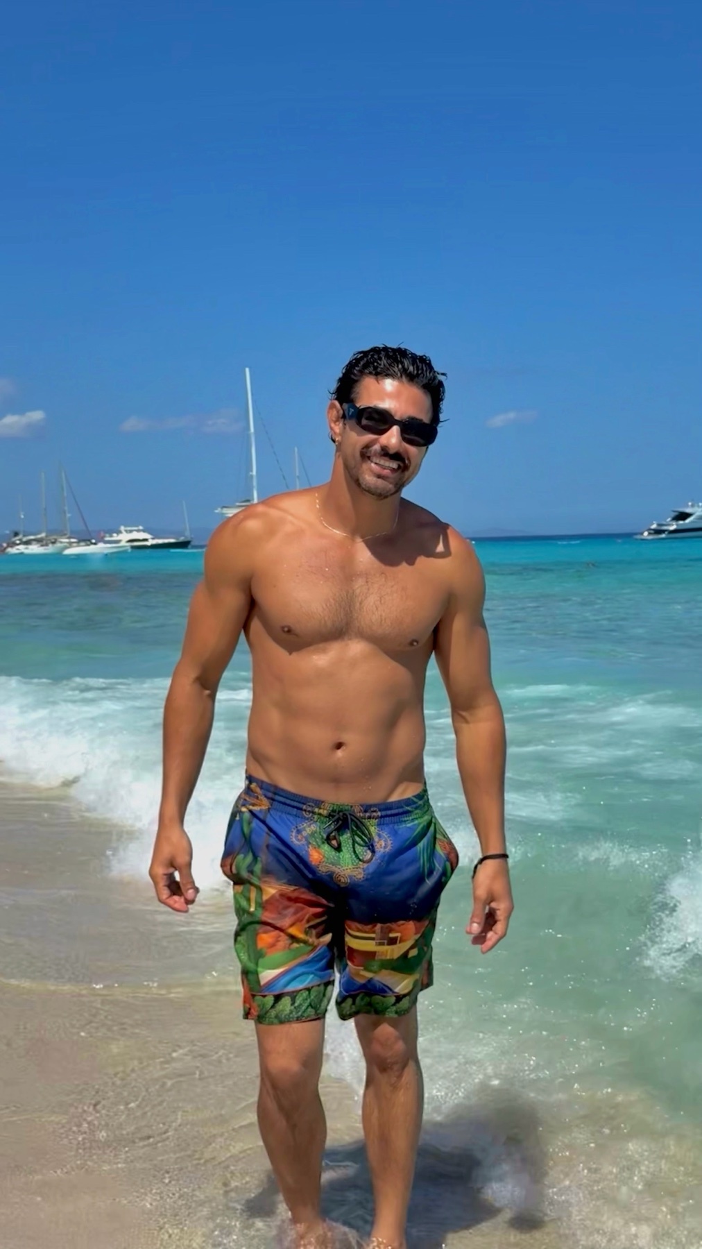 A man stands on a sandy beach wearing colorful swim trunks and sunglasses. He is shirtless, with the ocean and sailboats in the background under a clear blue sky.