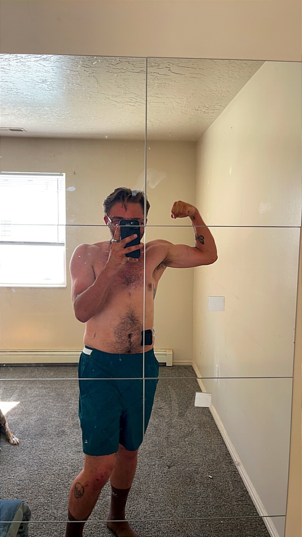 A man with glasses and tattoos flexes his bicep in a mirror selfie. He is shirtless and wearing shorts, with a dog visible in the background.