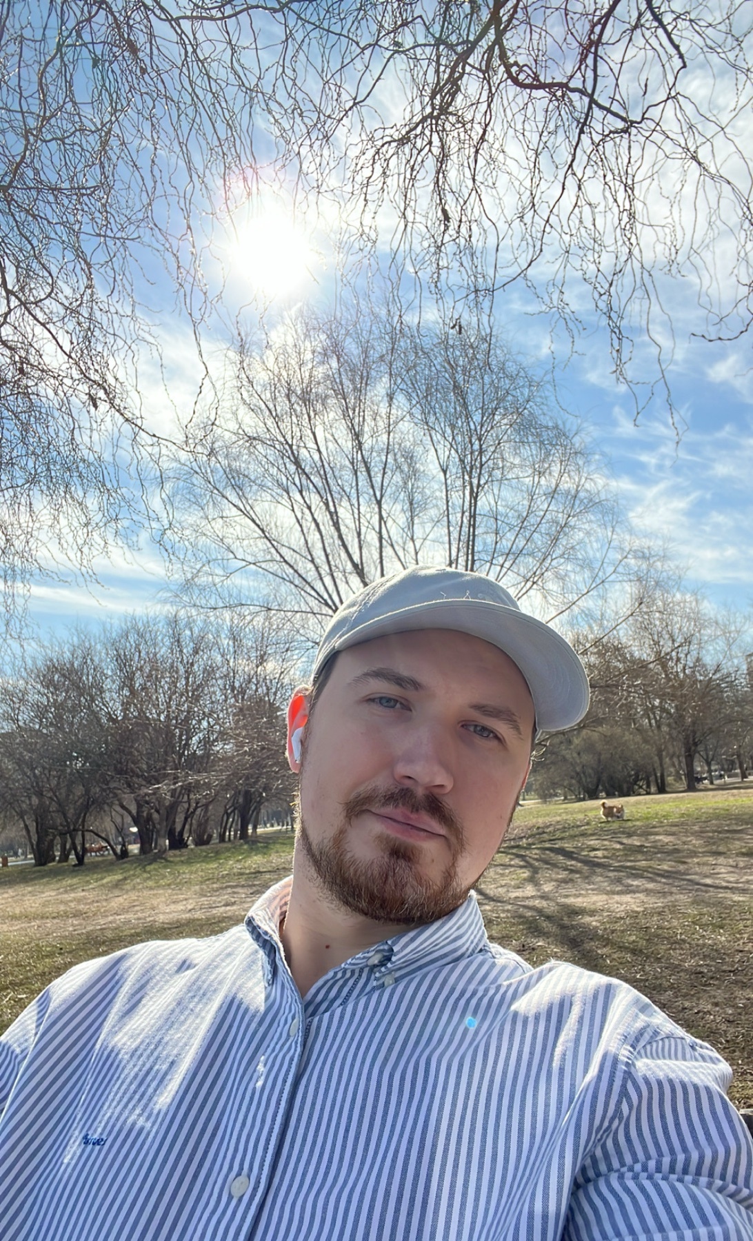 A man takes a selfie outdoors on a sunny day. He is wearing a cap and a striped shirt.