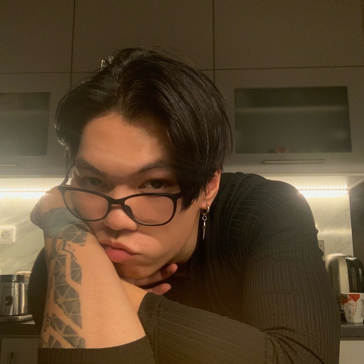 A man with black hair and glasses poses for a selfie. He is leaning on his arms in a kitchen setting.