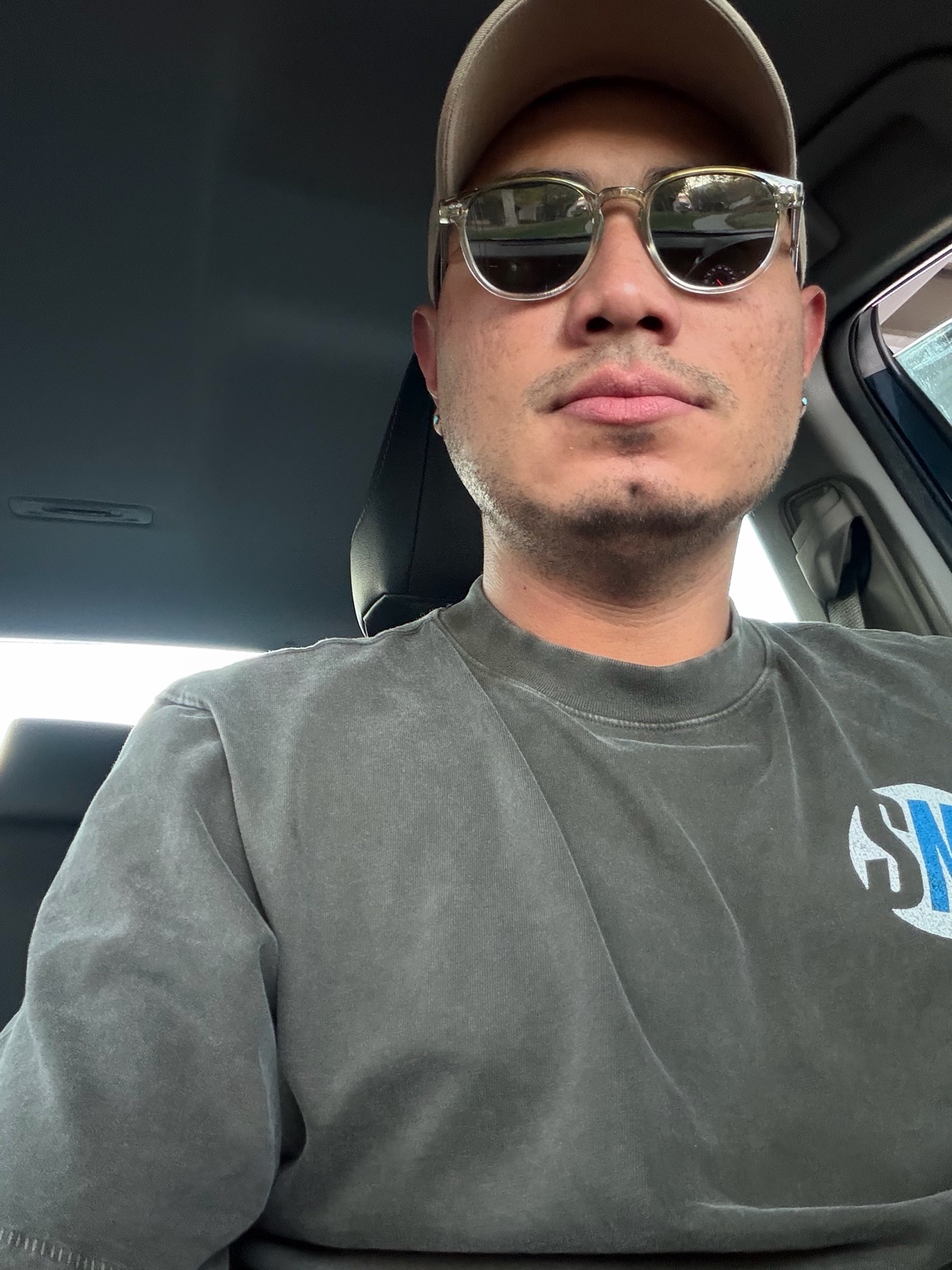 A man wearing sunglasses and a baseball cap takes a selfie in his car. He has stubble and a mustache.