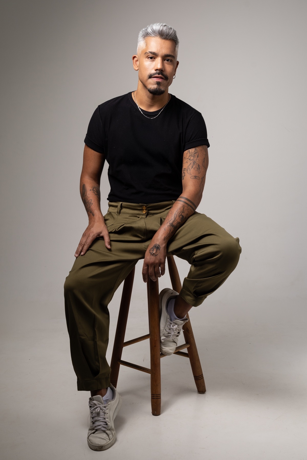 A man with grey-streaked hair and a moustache sits on a stool in a studio. He is wearing a black t-shirt, olive green pants, and white sneakers.