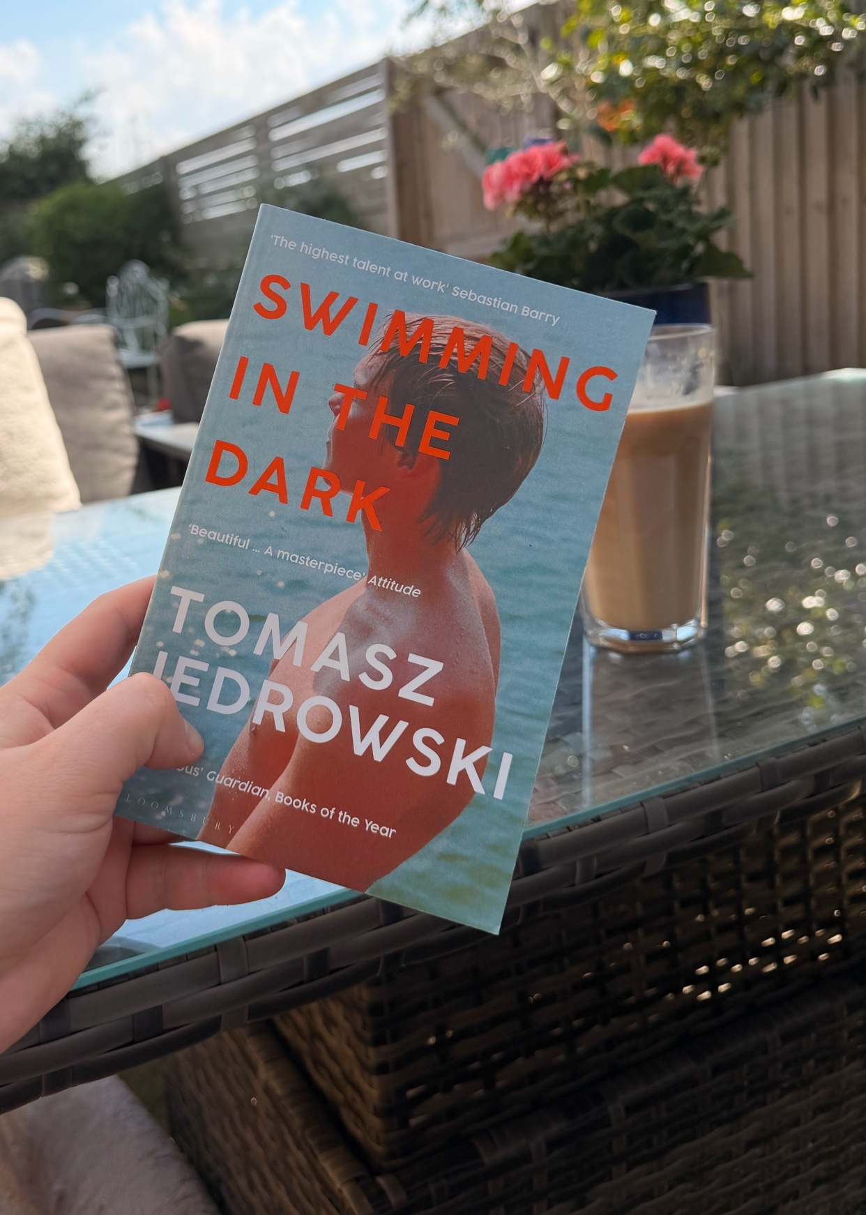 A hand holds a book titled "Swimming in the Dark" by Tomasz Jedrowski, with a glass of iced coffee and a garden setting in the background.