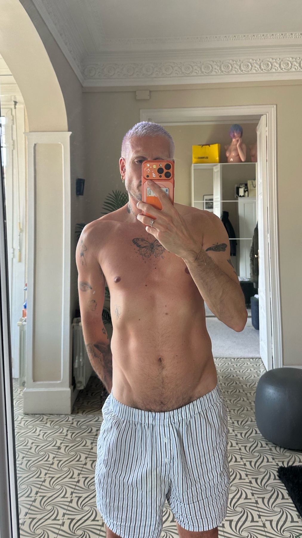 A man with purple hair takes a selfie in striped boxer briefs. He has tattoos on his arms and chest.