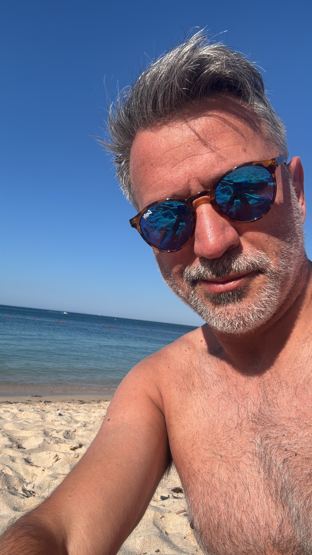 A man with grey hair and stubble, wearing sunglasses, takes a selfie on a sunny beach with the ocean and sand in the background.
