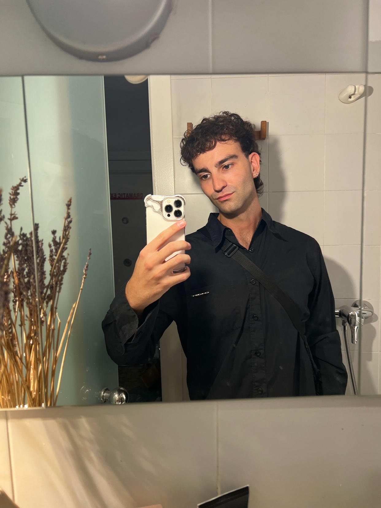 A man takes a mirror selfie in a bathroom. He is wearing a black shirt and has curly hair.