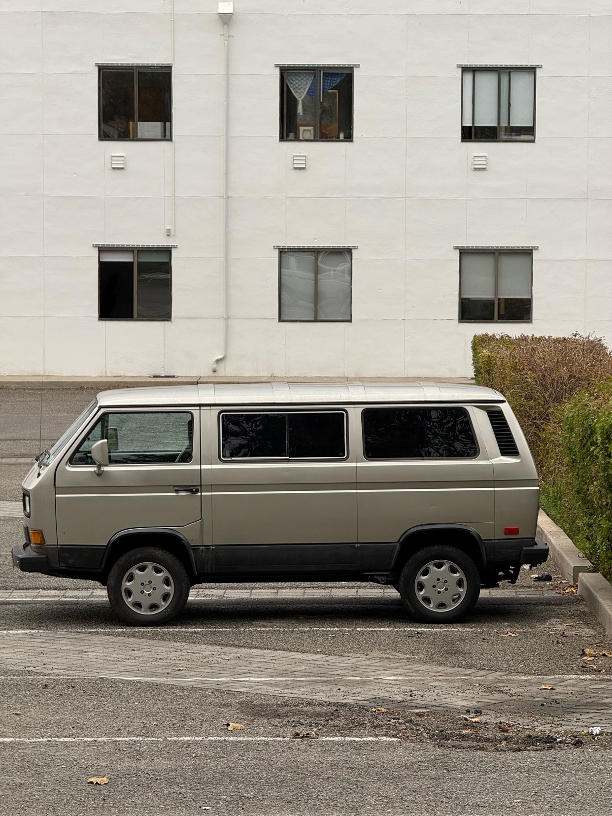A vintage Volkswagen van is parked in front of a white building. The van is a light grey color and appears to be in good condition.