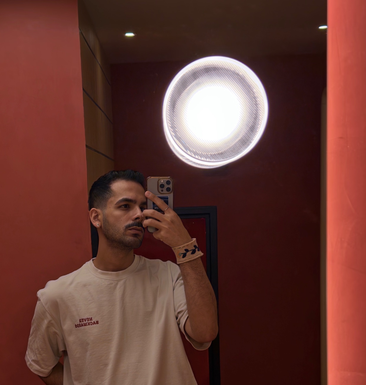 A man takes a selfie in a mirror, wearing a light-colored t-shirt and a bracelet. The background is a deep red wall with a circular light fixture reflecting in the mirror.