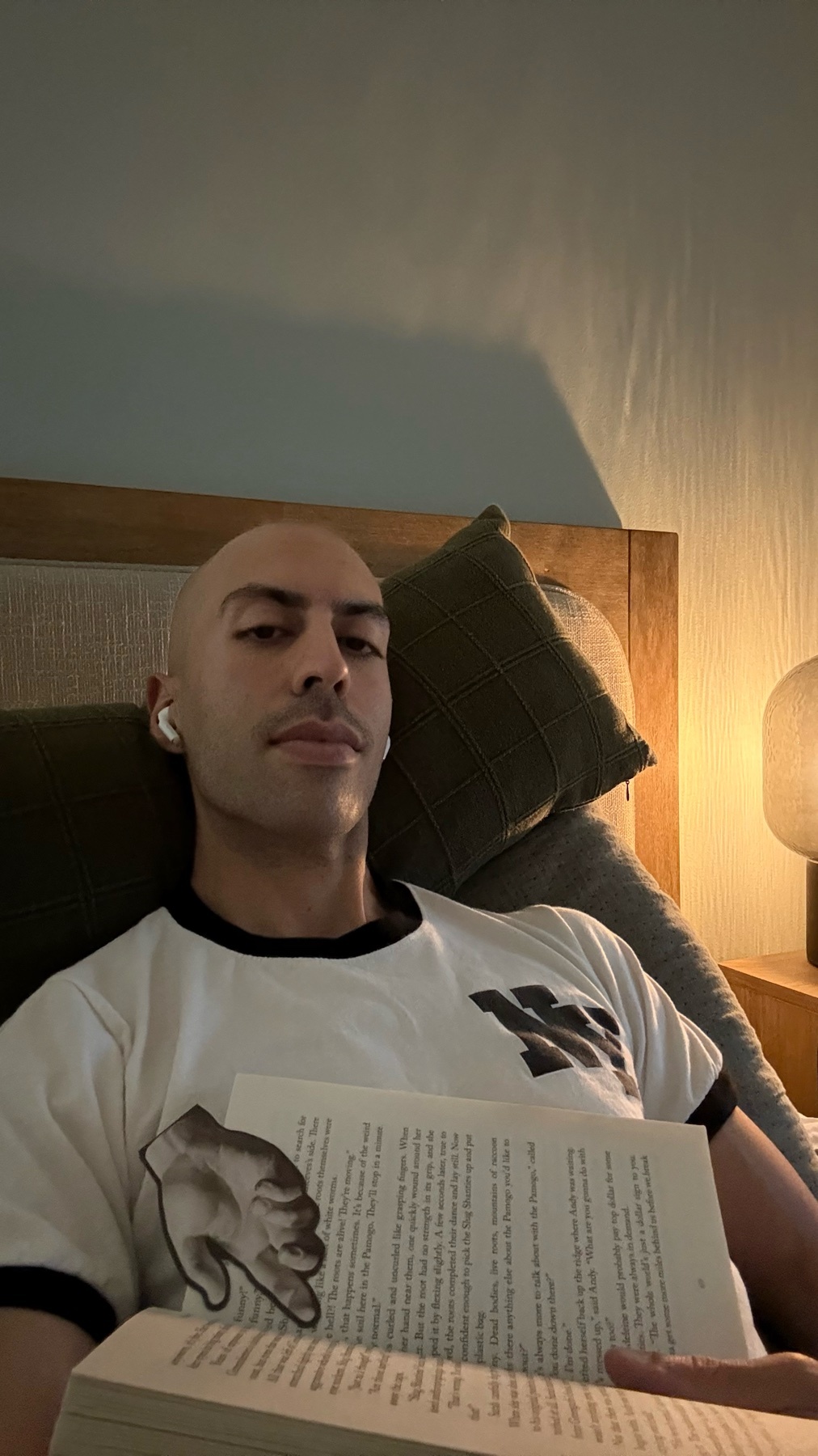 A bald man with earbuds is lying in bed, holding an open book. He is wearing a t-shirt with a graphic design on it.