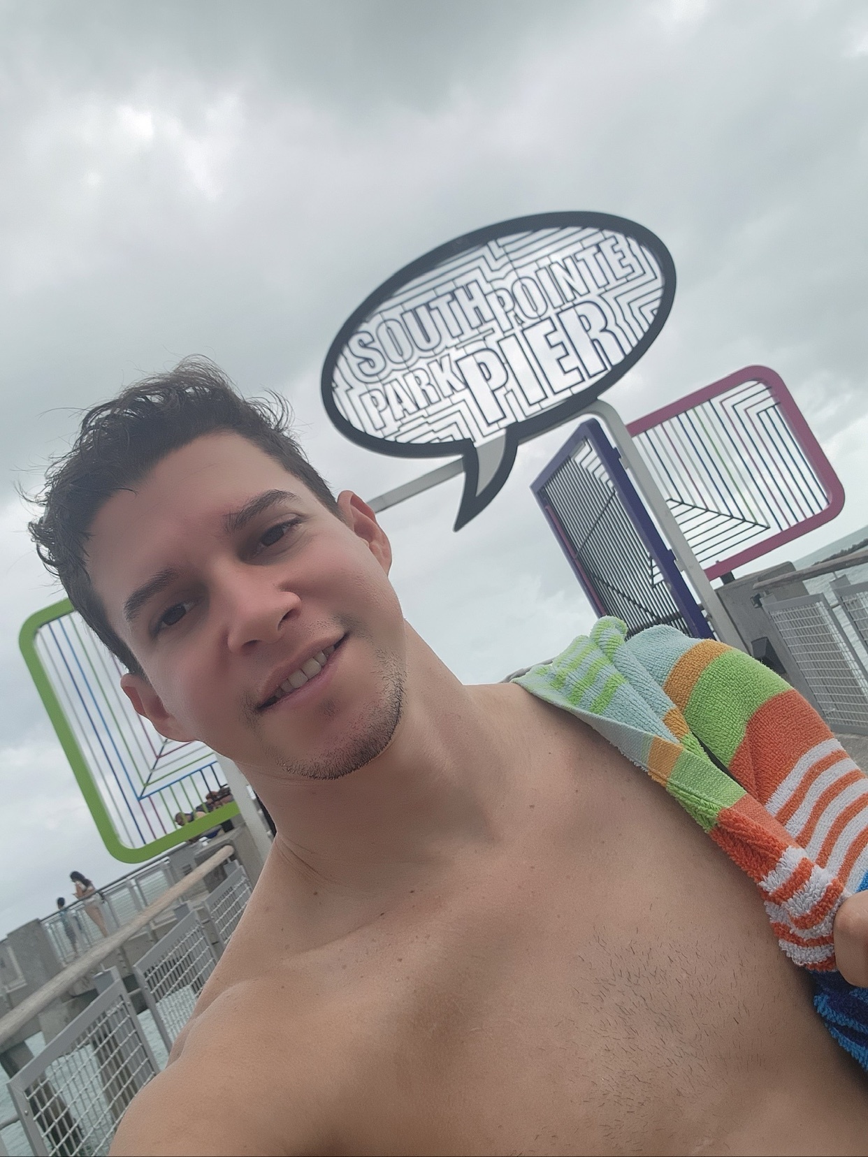 A man takes a selfie at South Pointe Park Pier. He is shirtless and smiling, with a towel over his shoulder.
