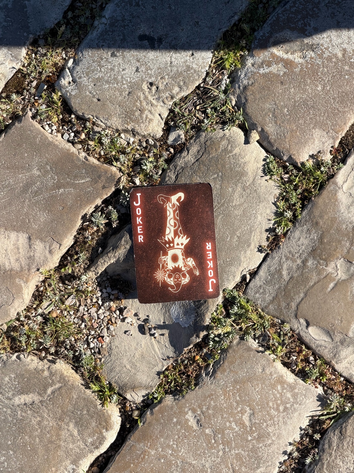 A single joker playing card lies face up on a cobblestone path, surrounded by patches of grass and weeds.