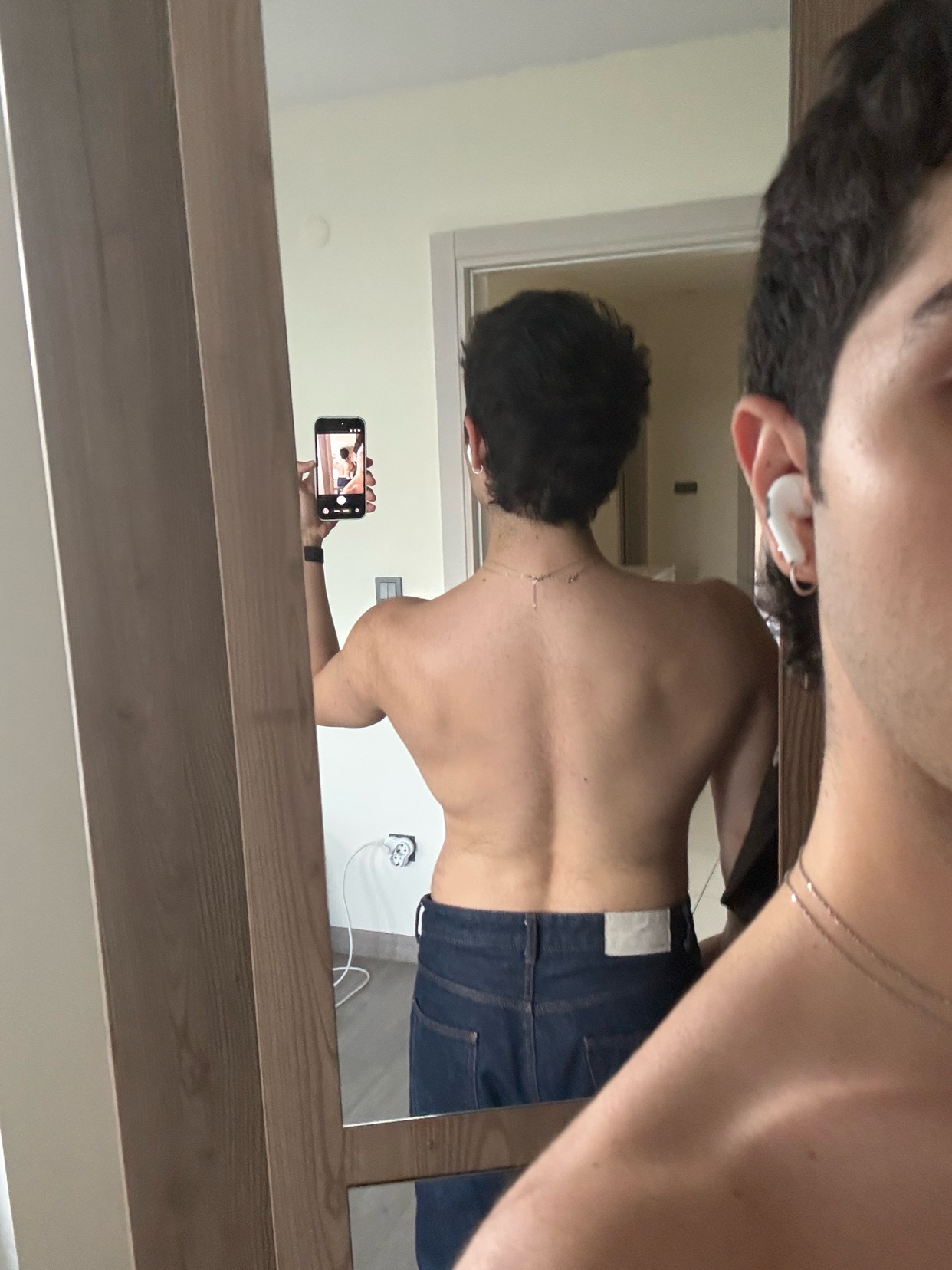 A man takes a mirror selfie, showcasing his back and physique. He is shirtless, wearing jeans, and has dark hair.