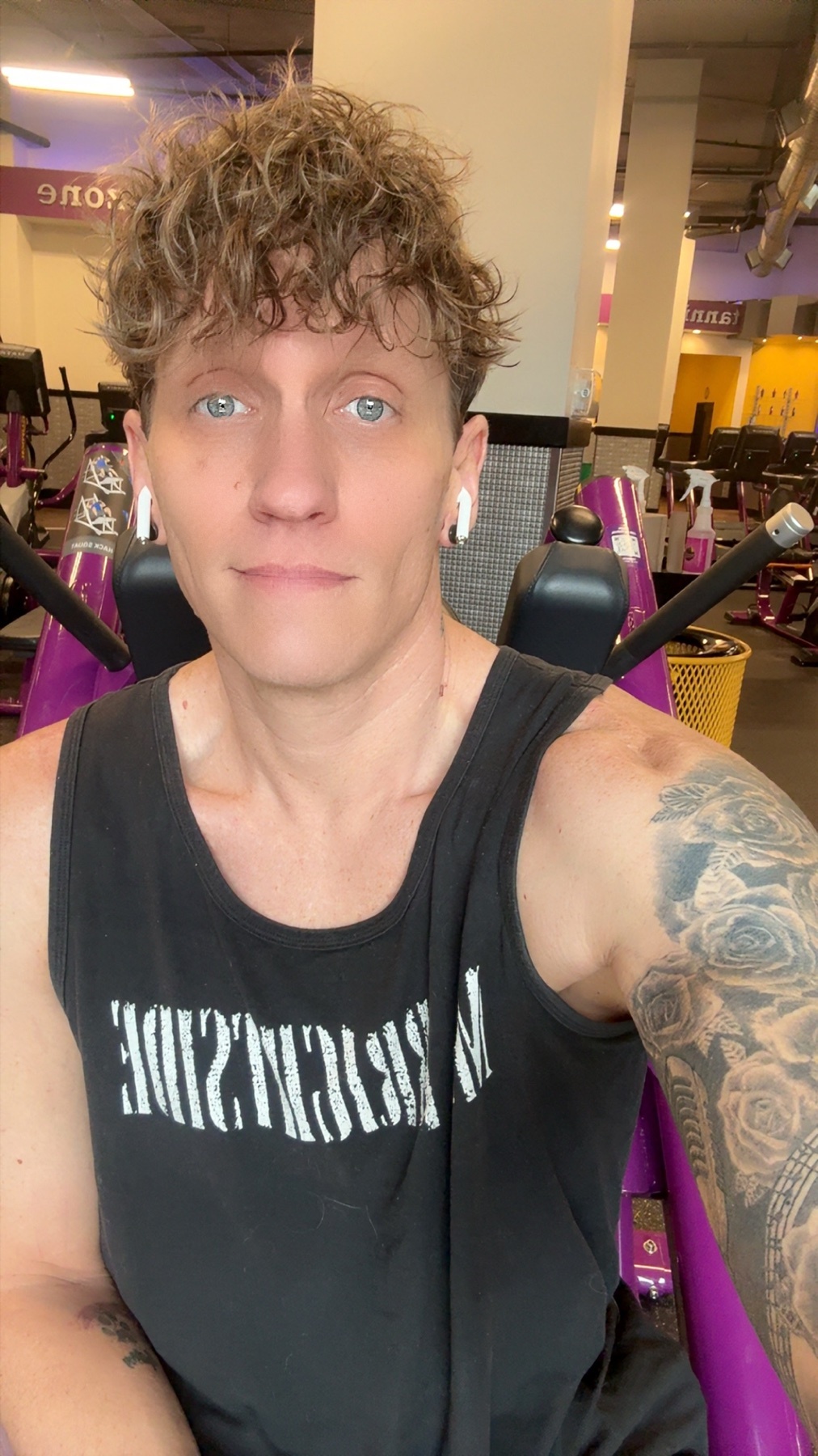A person with curly hair and tattoos is taking a selfie at the gym. They are wearing earbuds and a black tank top.