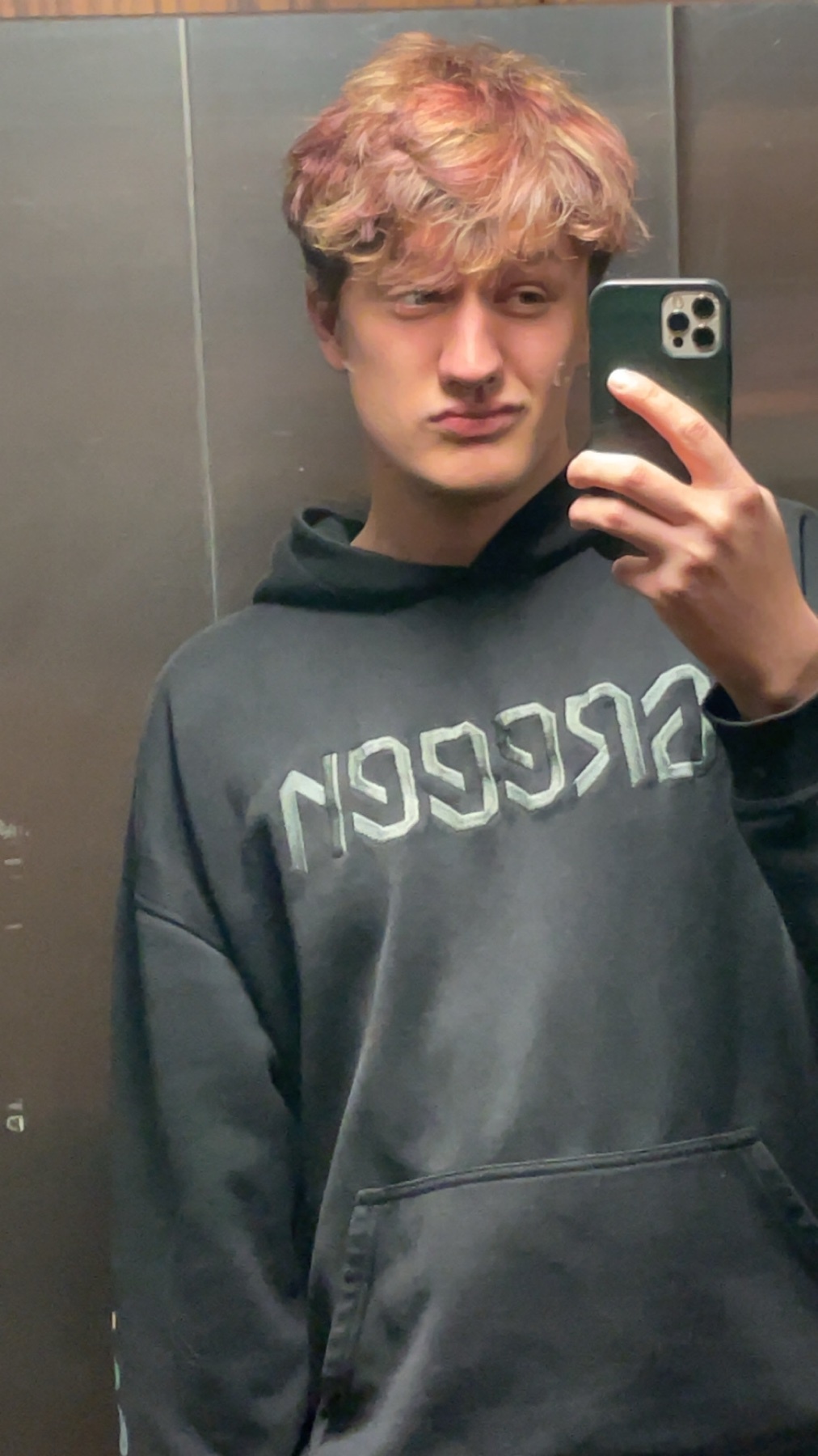 A young man takes a selfie in an elevator. He is wearing a black hoodie and has curly, dark hair.
