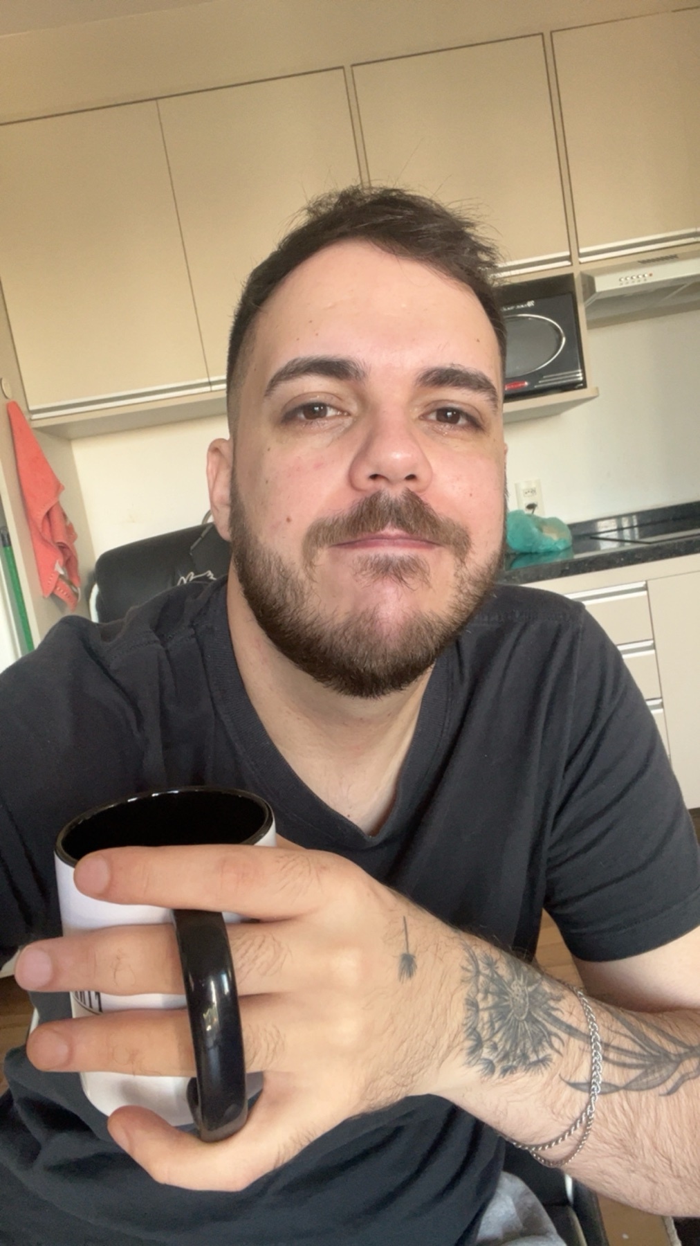 A man with a beard and tattoos holds a coffee mug while looking at the camera.
