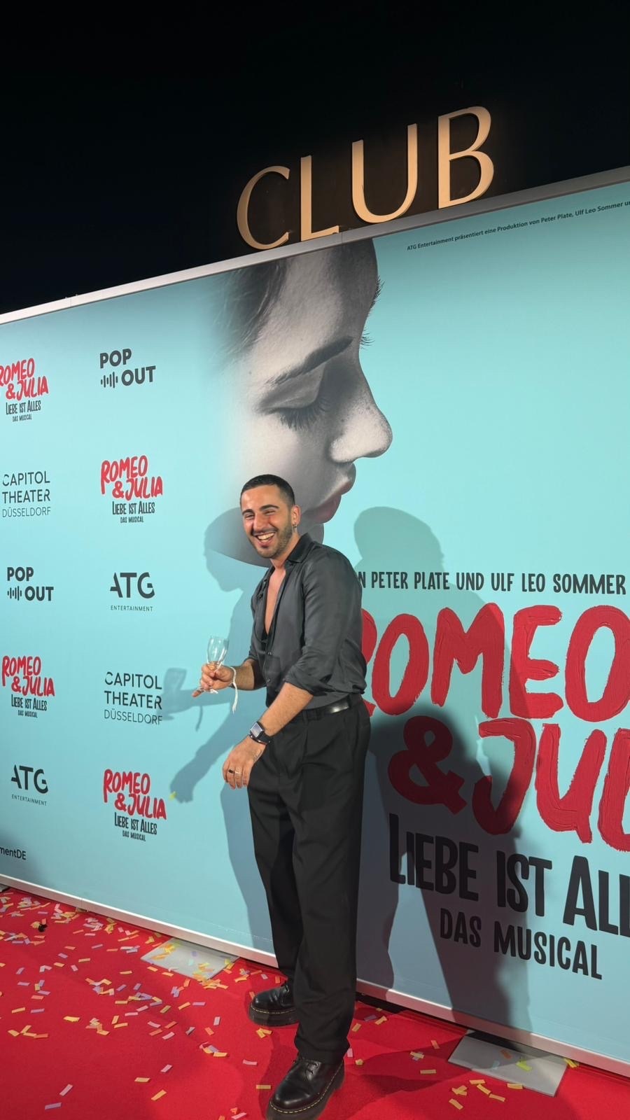 A man in a dark suit and open-collared shirt poses on a red carpet in front of a backdrop for the musical "Romeo & Julia". He holds a glass of champagne and smiles at the camera.