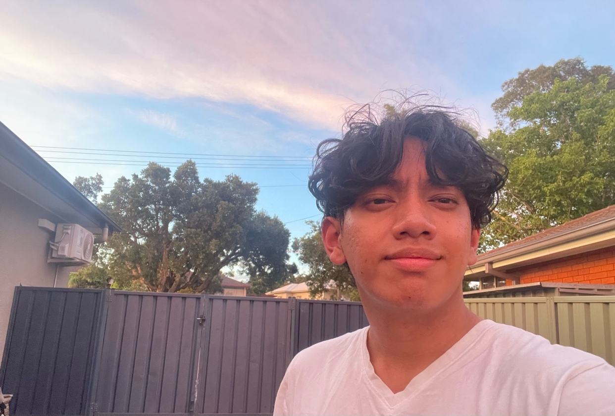 A young man takes a selfie outdoors. He is wearing a white t-shirt and has curly dark hair.