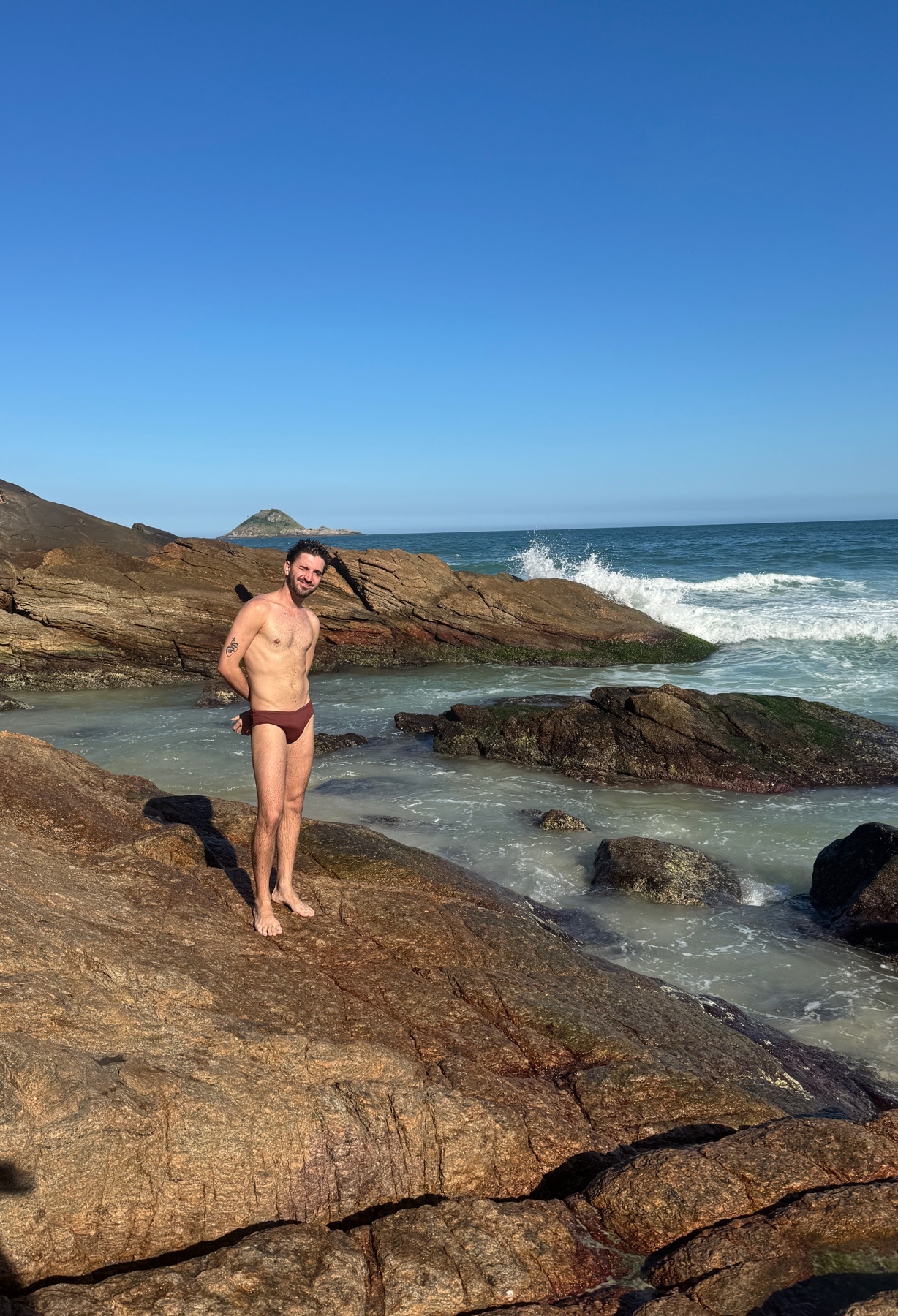 A man stands on a rocky beach wearing dark red swim briefs. The ocean waves crash against the rocks behind him under a clear blue sky.
