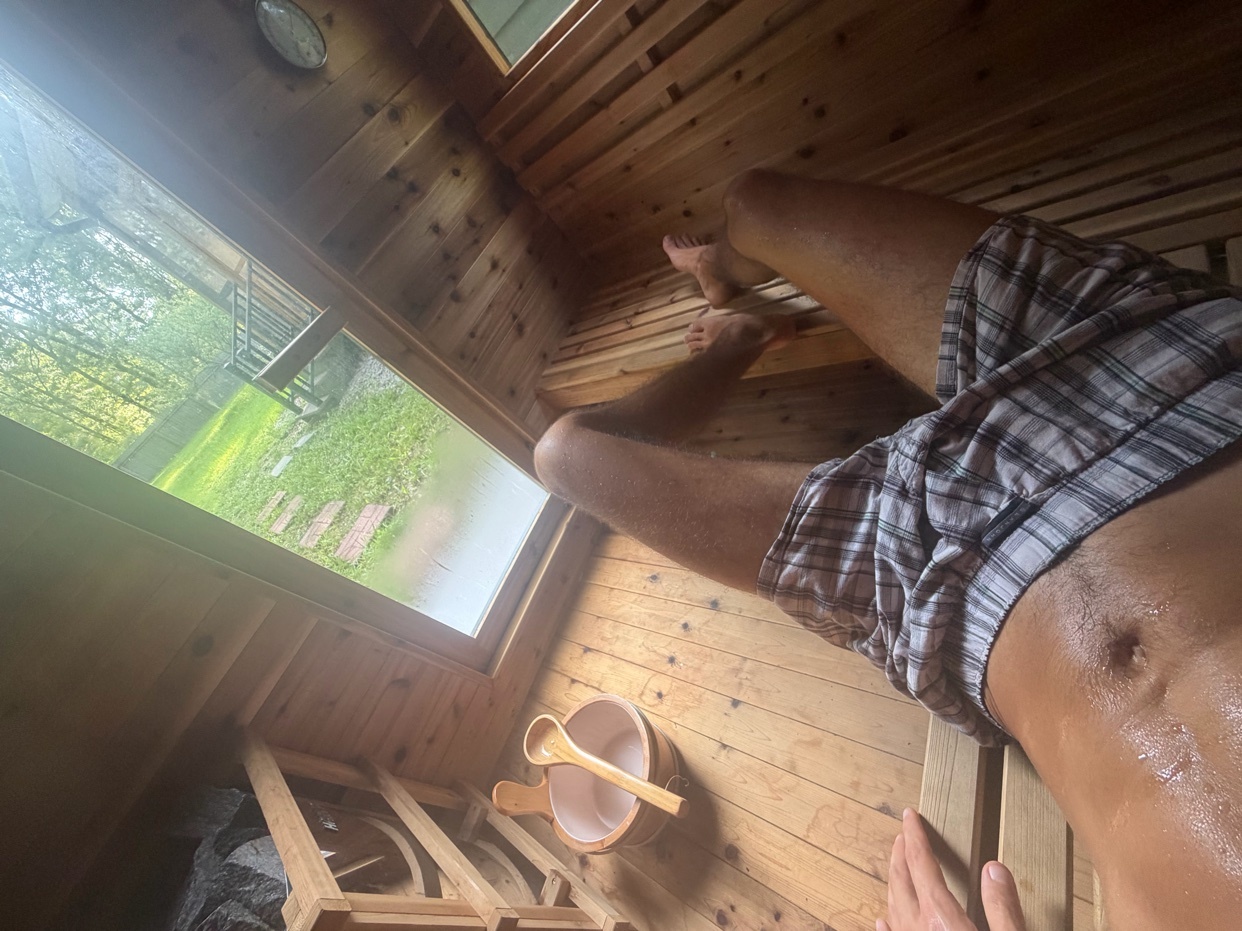 A man relaxes in a wooden sauna, glistening with sweat. Outside the window, a lush green yard is visible.