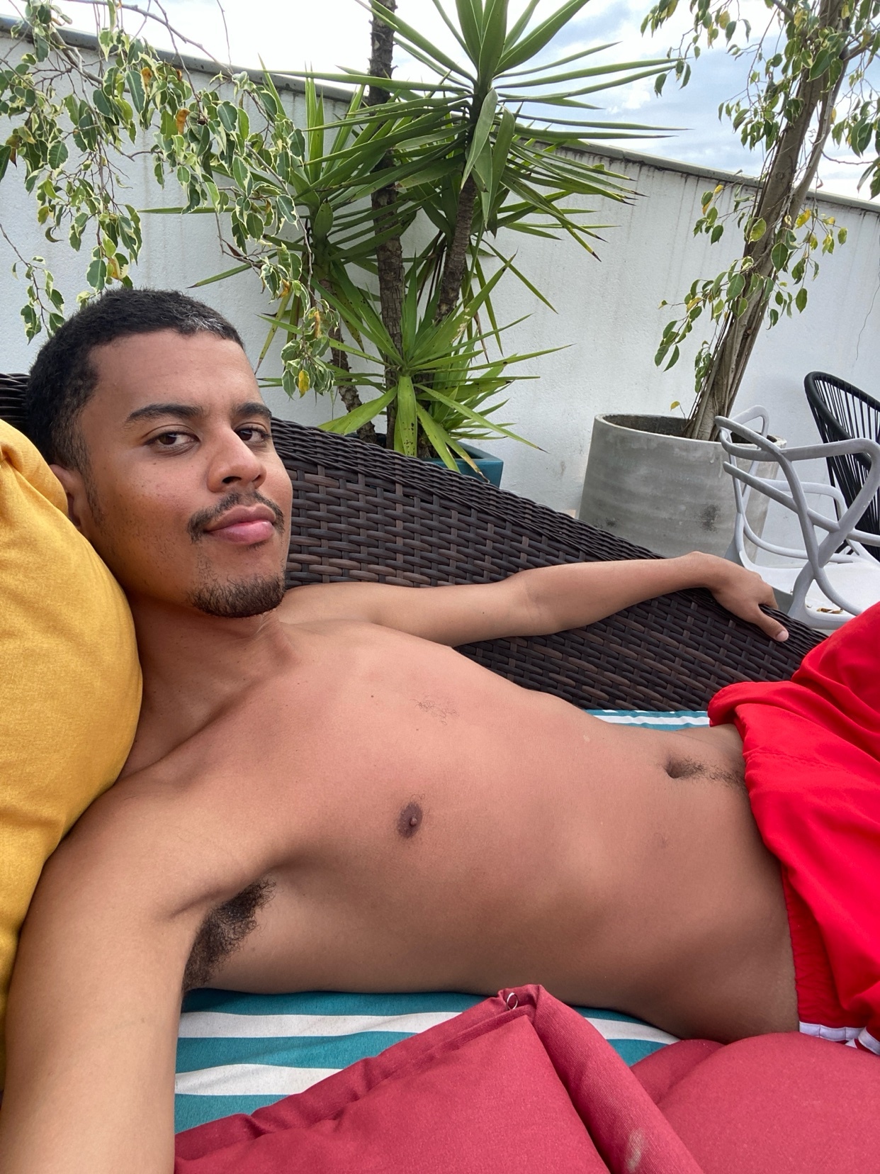 A shirtless man poses on a patio. He is wearing red shorts and has dark hair and stubble.