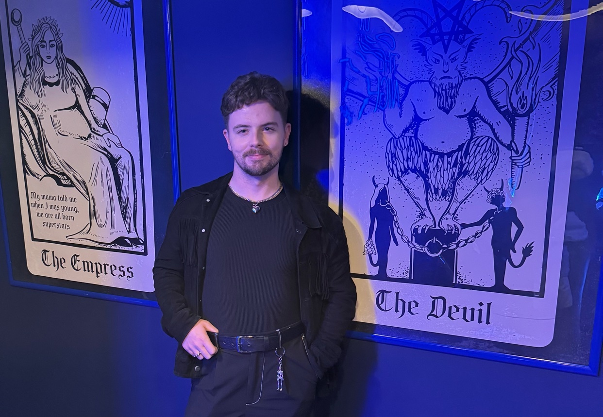 A man poses in front of two tarot cards, "The Empress" and "The Devil". The image has a dark, modern aesthetic.
