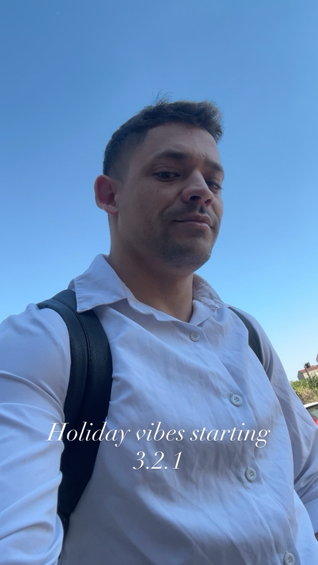 A man in a white button-down shirt looks up at the camera with a backpack on. The text "Holiday vibes starting 3.2.1" is overlaid on the image.