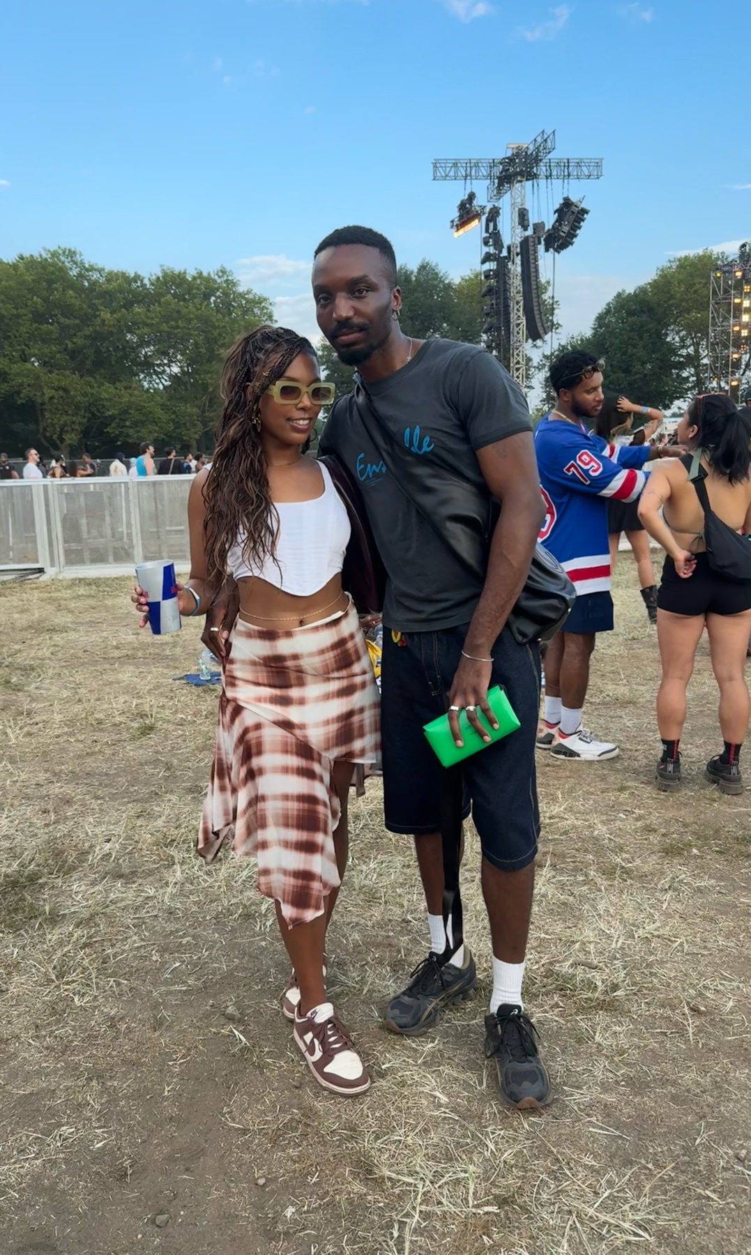 A couple poses for a photo at an outdoor festival. The woman wears a white top and a patterned skirt, while the man wears a t-shirt and shorts.