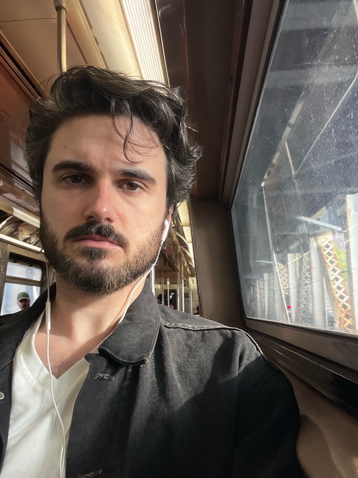 A man with dark hair and a beard takes a selfie on a train, wearing earbuds and a dark jacket over a white t-shirt.
