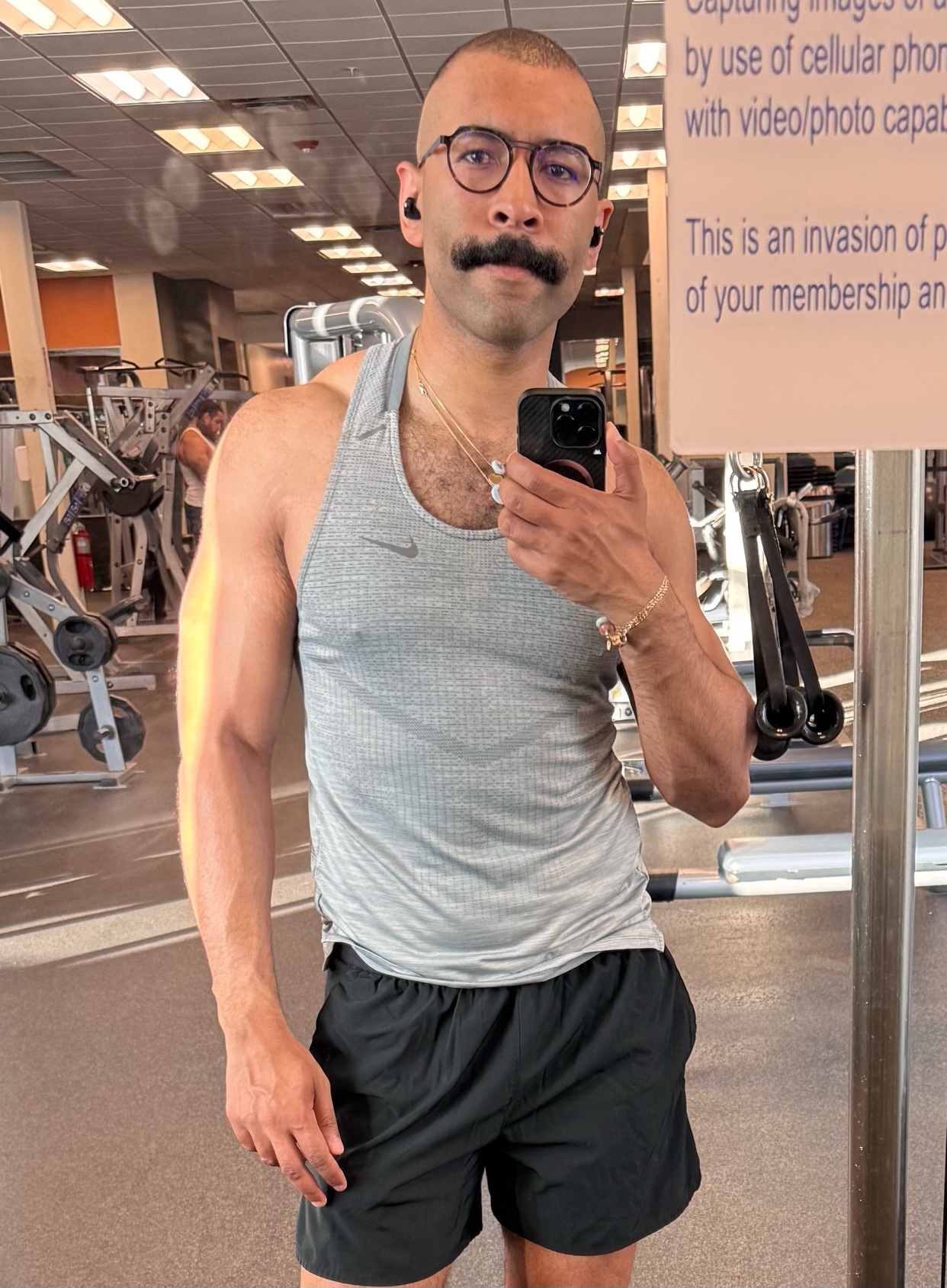 A man with a mustache and glasses takes a selfie in a gym, wearing a tank top and shorts. He is flexing his bicep and has earbuds in.