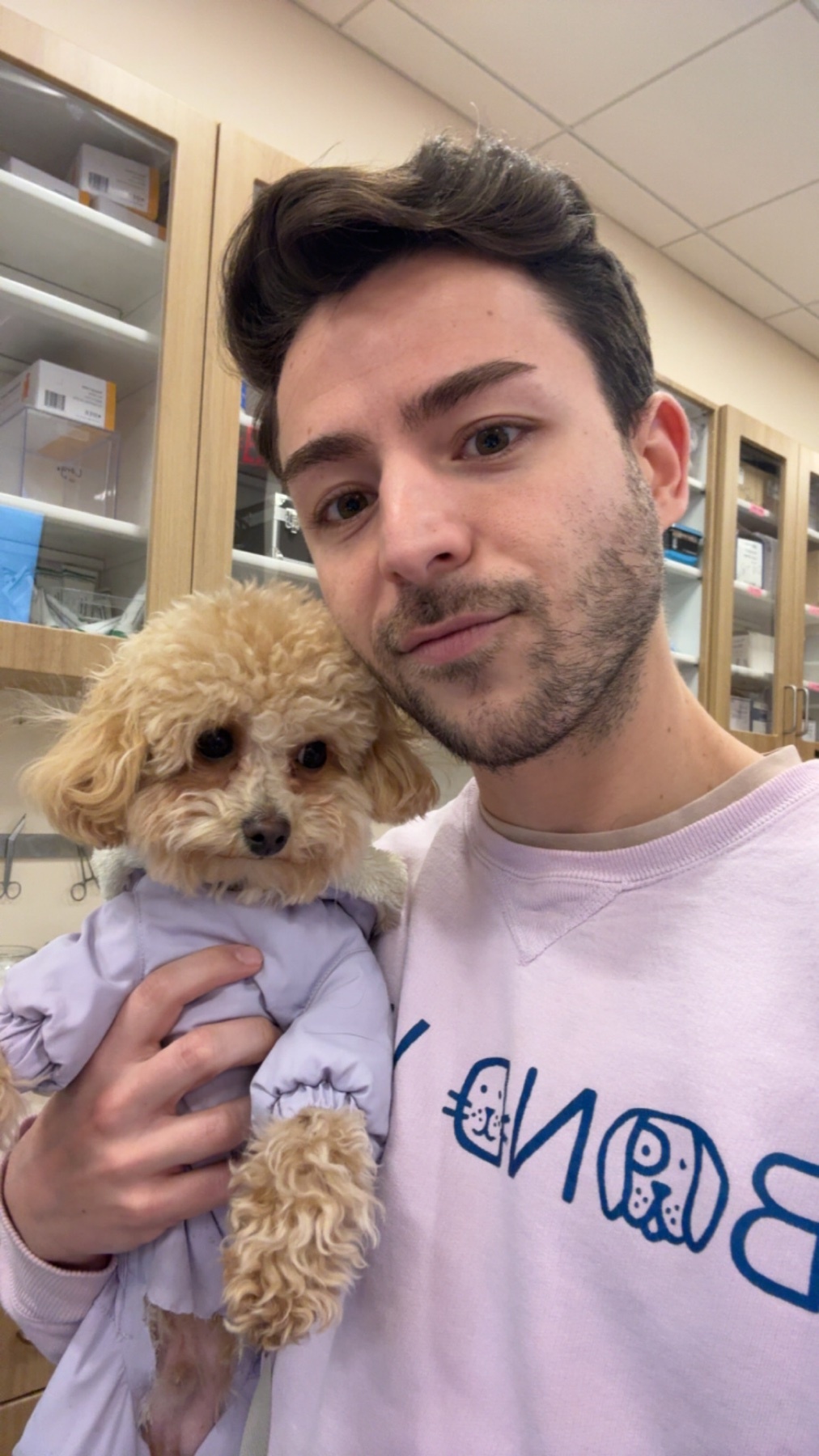A man takes a selfie while holding a small dog. The man is smiling and the dog is wearing a purple outfit.