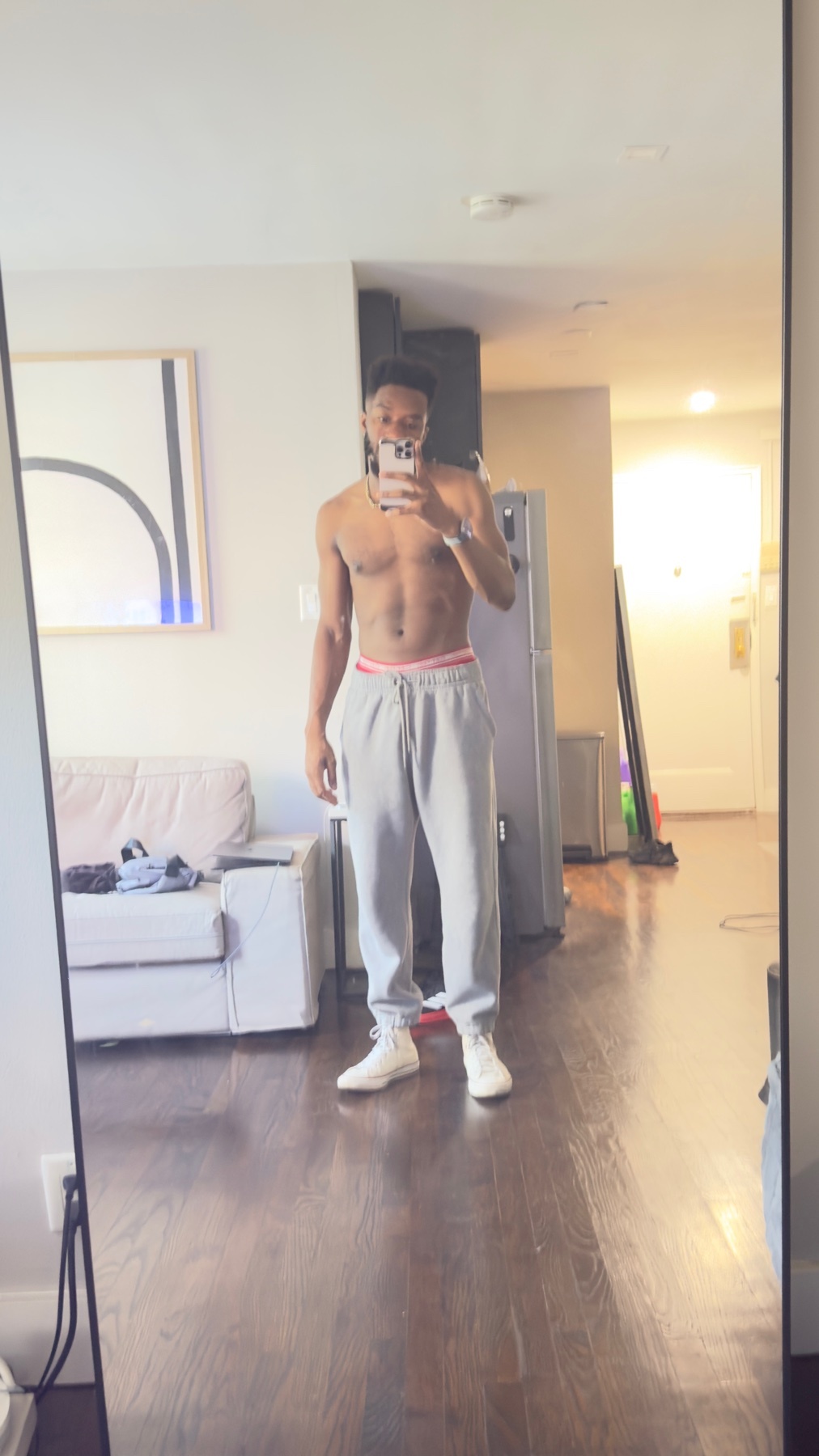 A man takes a mirror selfie, shirtless, showcasing his physique. He's wearing gray sweatpants and white sneakers.