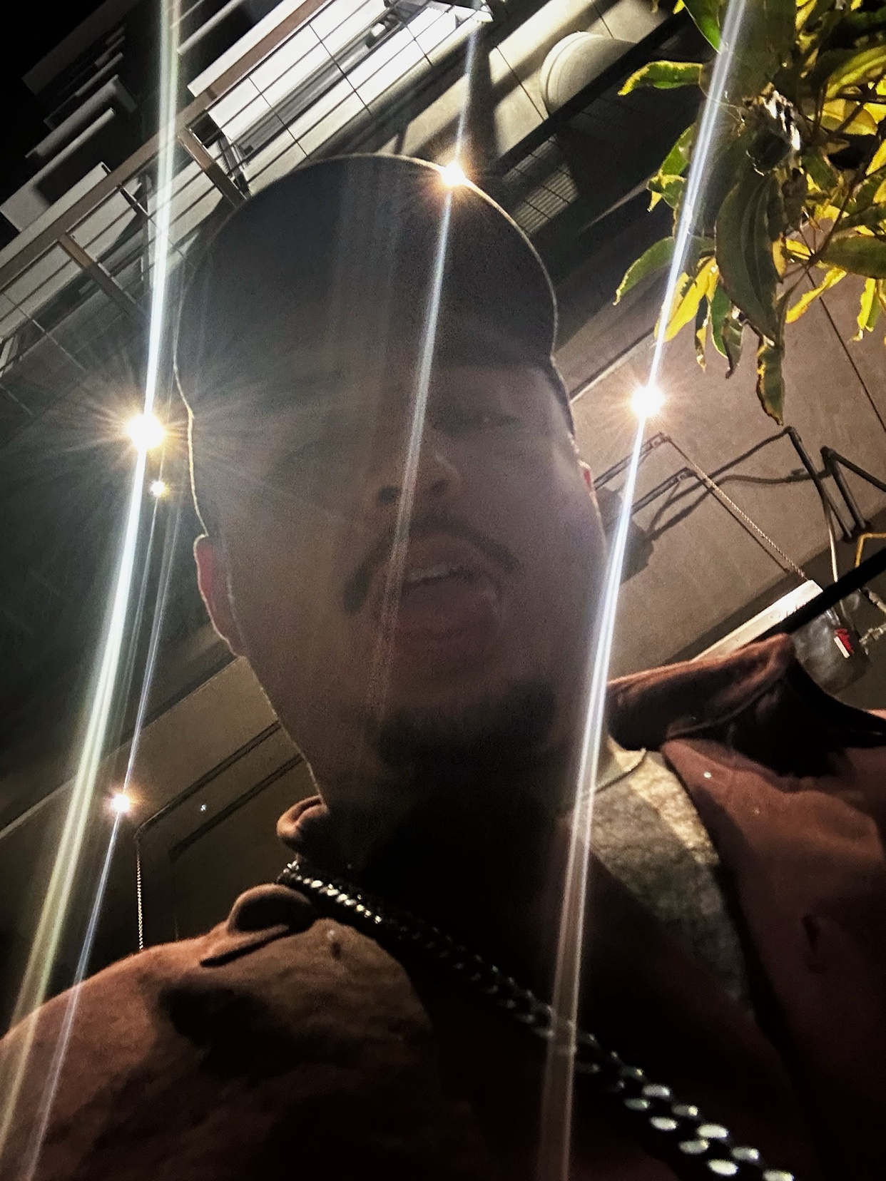 A man is shirtless, wearing a cap and chain. The photo is taken at night with bright lights.