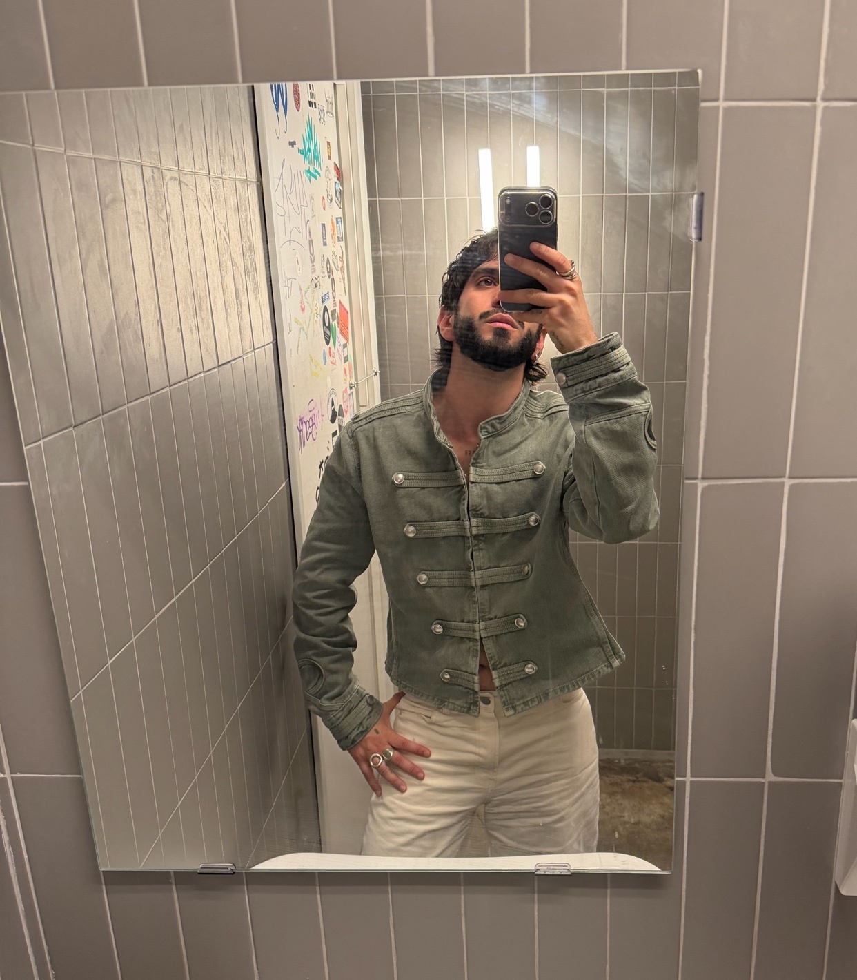 A man with a beard and dark hair takes a mirror selfie in a bathroom. He is wearing a green military-style jacket and white pants.