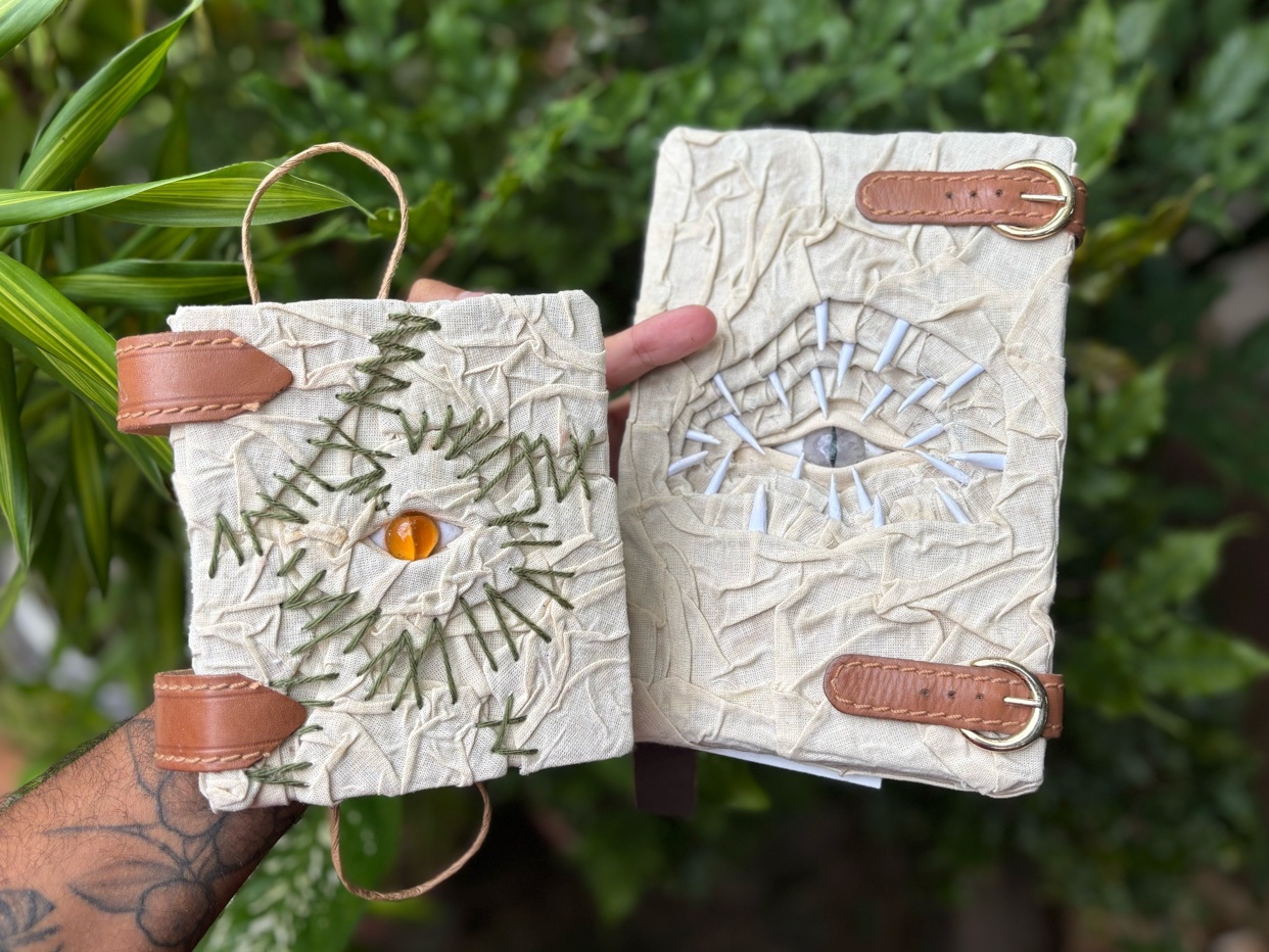 Two handmade journals with textured, off-white covers are displayed. The journal on the left features green embroidery resembling a starburst with an orange gem at its center, secured by leather straps and buckles. The journal on the right has an emb